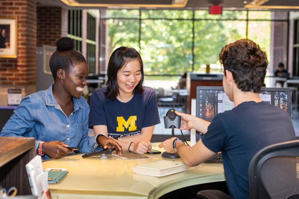 Three U-M students laugh as they gather around a computer workstation