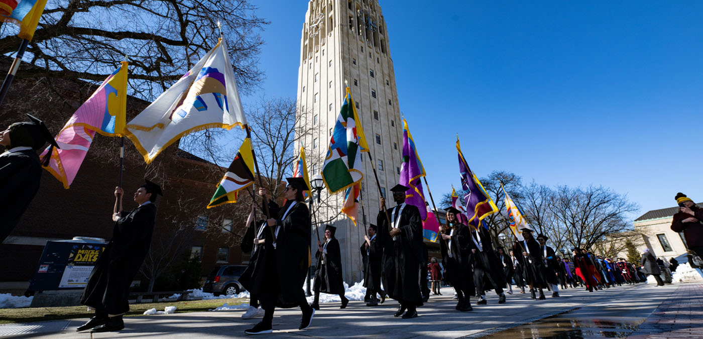 Graduates with flags walk past the Burton tower.